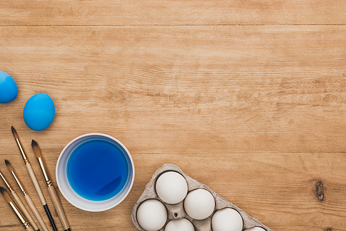 top view of watercolor blue paint in bowl near chicken eggs and paintbrushes on wooden table