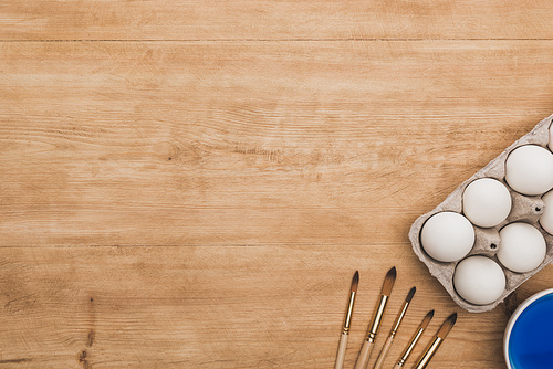 top view of watercolor blue paint in bowl near chicken eggs and paintbrushes on wooden table