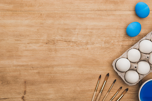 top view of watercolor blue paint in bowl near chicken eggs and paintbrushes on wooden table