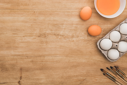 top view of watercolor orange paint in bowl near chicken eggs and paintbrushes on wooden table