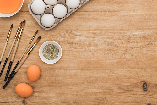 top view of orange watercolor paint and golden glitter in bowls near Easter eggs and paintbrushes on wooden table