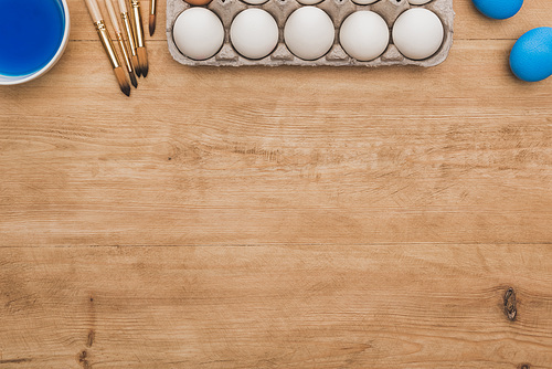 top view of blue watercolor paint in bowl near chicken eggs and paintbrushes on wooden table