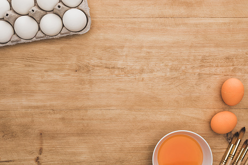 top view of orange watercolor paint in bowl near chicken eggs and paintbrushes on wooden table