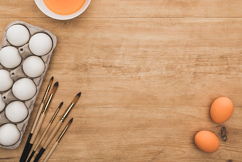 top view of orange watercolor paint in bowl near chicken eggs and paintbrushes on wooden table