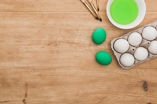 top view of green watercolor paint in bowl near chicken eggs and paintbrushes on wooden table