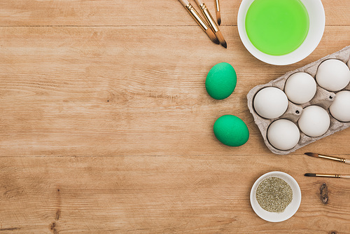 top view of green watercolor paint and glitter in bowls near Easter eggs and paintbrushes on wooden table