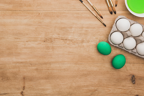 top view of green watercolor paint in bowl near chicken eggs and paintbrushes on wooden table