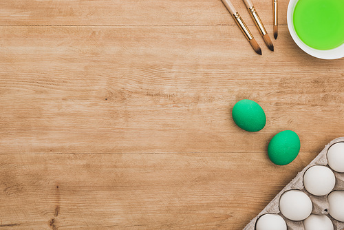 top view of green watercolor paint in bowl near chicken eggs and paintbrushes on wooden table