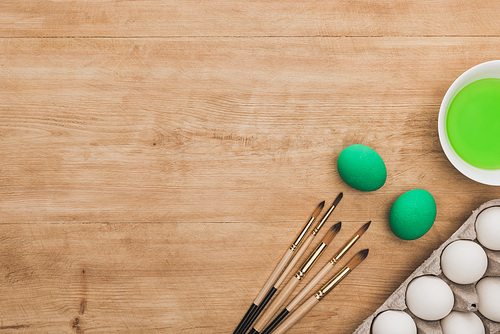 top view of green watercolor paint in bowl near chicken eggs and paintbrushes on wooden table