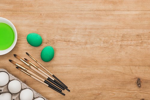 top view of green watercolor paint in bowl near chicken eggs and paintbrushes on wooden table