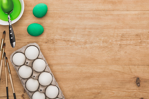 top view of green watercolor paint in bowl near chicken eggs and paintbrushes on wooden table