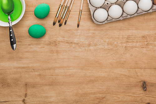 top view of green watercolor paint in bowl near chicken eggs and paintbrushes on wooden table