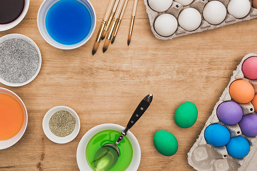 top view of watercolor paints and glitter in bowls near Easter eggs and paintbrushes on wooden table