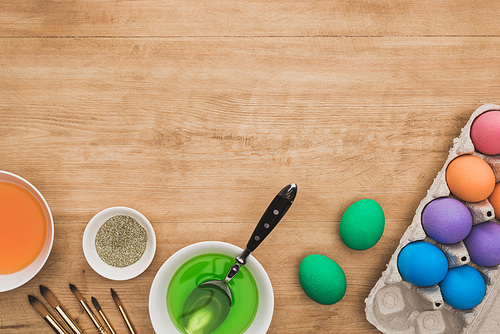 top view of watercolor paints and glitter in bowls near Easter eggs and paintbrushes on wooden table