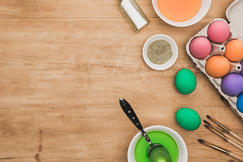 top view of watercolor paints and glitter in bowls near Easter eggs and paintbrushes on wooden table