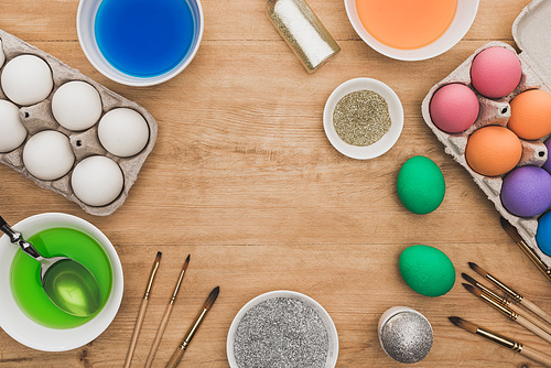 top view of watercolor paints and glitter in bowls near Easter eggs and paintbrushes on wooden table