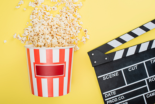 top view of bucket with tasty popcorn near clapperboard on yellow, cinema concept