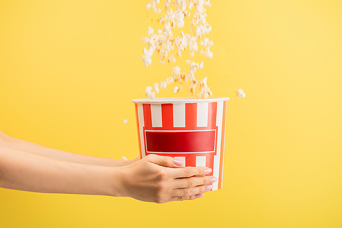 tasty popcorn falling into striped bucket in female hands isolated on yellow, cinema concept