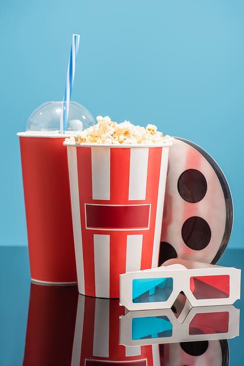 cup of soda, bucket with popcorn, 3d glasses and film bobbin on glossy surface isolated on blue, cinema concept