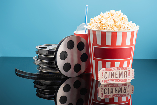 film reels, cup of soda, cinema ticket and bucket of popcorn on glossy surface isolated on blue