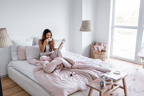Woman drinking coffee and reading book in modern bedroom
