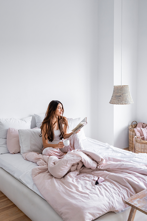 Smiling woman with coffee cup holding book on bed