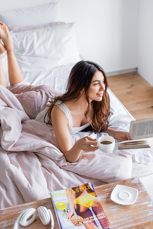 Smiling woman relaxing with coffee and book on bed