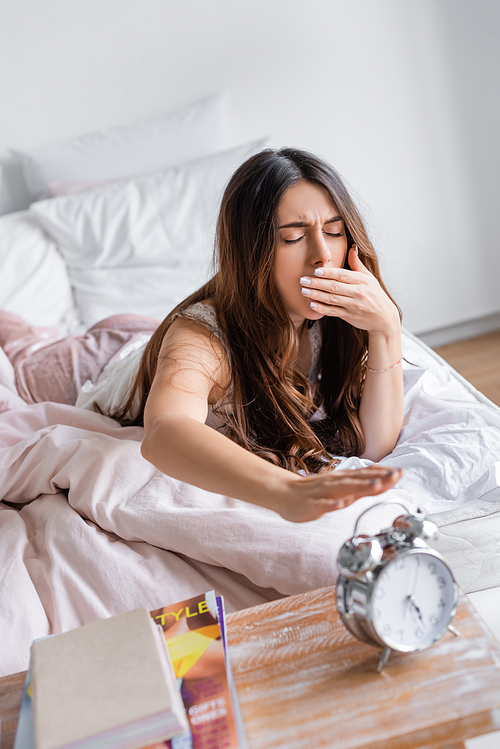 Yawning woman pulling hand to alarm clock on blurred foreground in bedroom