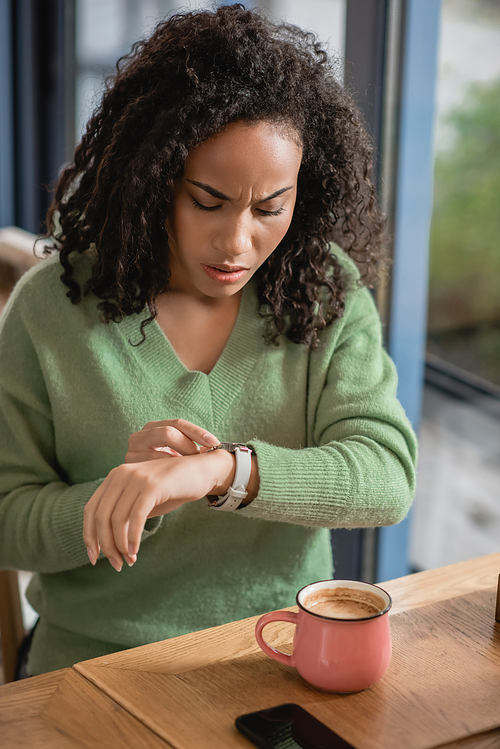 worried african american woman checking watch on wrist while waiting in cafe