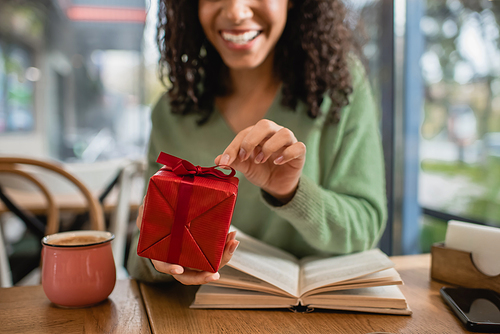 cropped view of happy african american woman pulling ribbon on red christmas gift box in cafe
