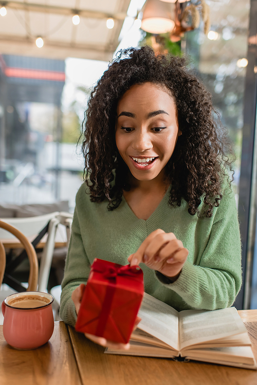 happy african american woman pulling ribbon on red christmas gift box in cafe