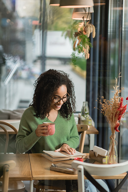 african american woman in eyeglasses holding cup of coffee and reading book in cafe
