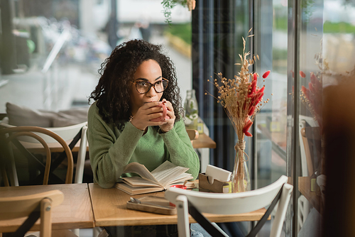 african american woman in eyeglasses drinking coffee near book on table in cafe