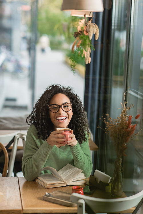amazed african american woman in eyeglasses holding cup of coffee near book on table in cafe