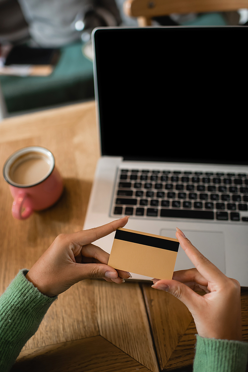 cropped view of african american woman holding credit card near laptop with blank screen