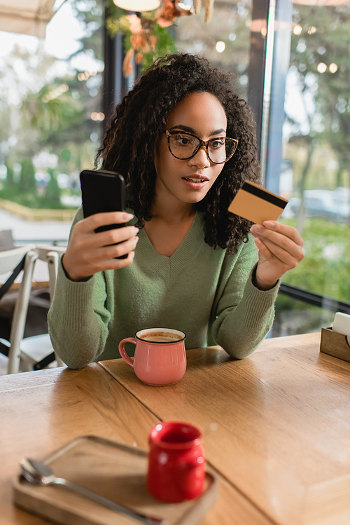african american woman looking at credit card while holding smartphone in cafe