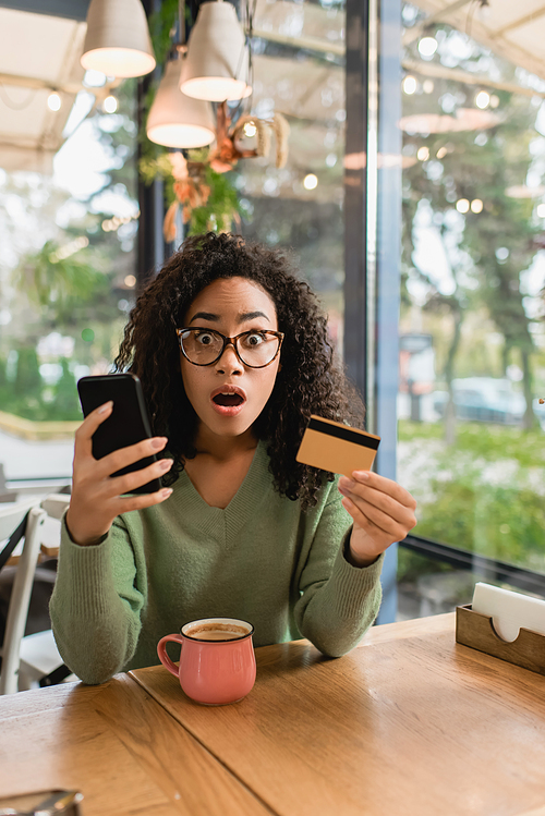 shocked african american woman holding smartphone and credit card in cafe