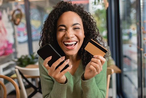 cheerful african american woman laughing while holding smartphone and credit card