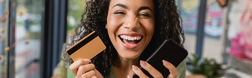 cheerful african american woman laughing while holding smartphone and credit card, banner