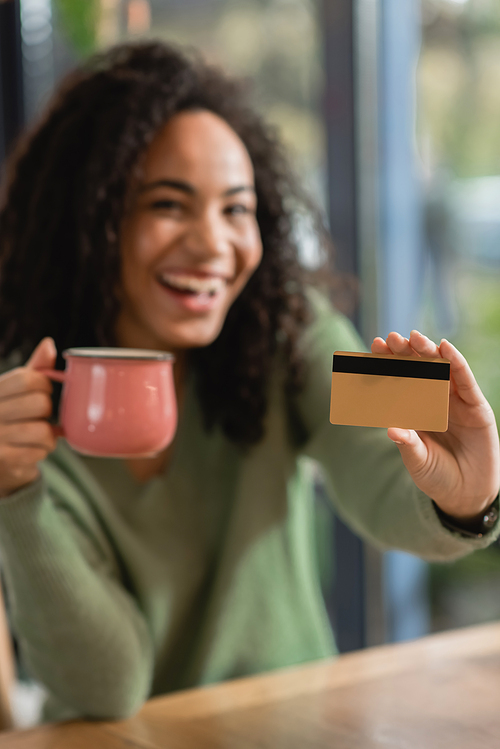 credit card and cup in hands of cheerful african american woman on blurred background