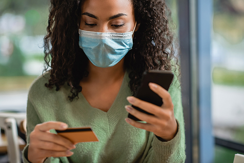 curly african american woman in medical mask holding smartphone and credit card in cafe