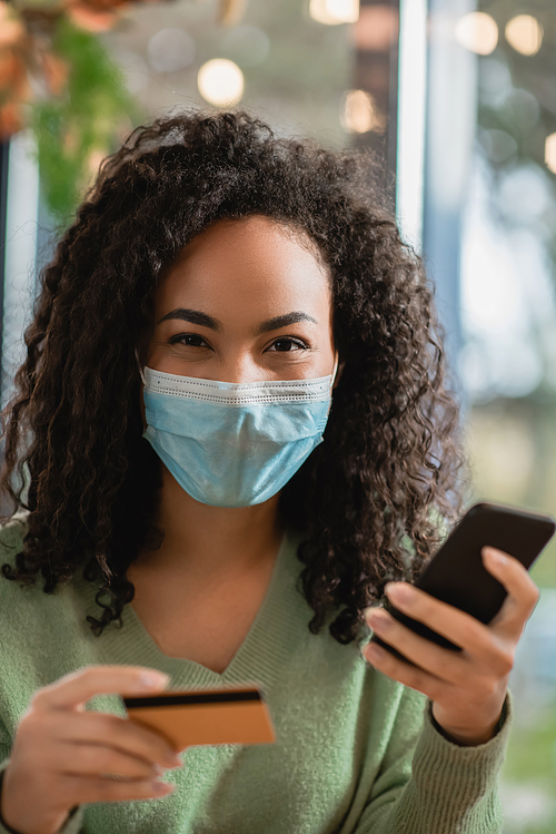 african american woman in medical mask holding smartphone and credit card in cafe