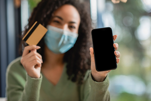 smartphone with blank screen and credit card in hands of african american woman in medical mask on blurred background
