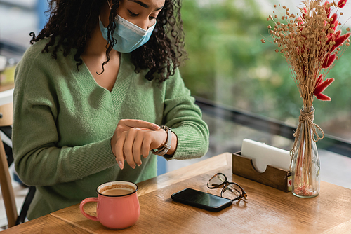 african american woman in medical mask looking at watch near smartphone with blank screen and cup of coffee on table