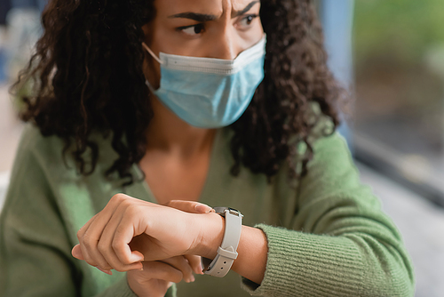 displeased african american woman in medical mask touching watch on wrist while waiting in cafe