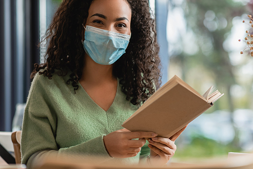african american woman in medical mask holding book in cafe