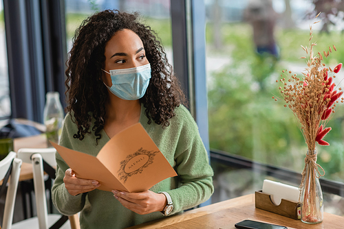 african american woman in medical mask holding menu and looking away in cafe