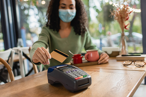 credit card near credit card reader in hand of african american woman in medical mask on blurred background
