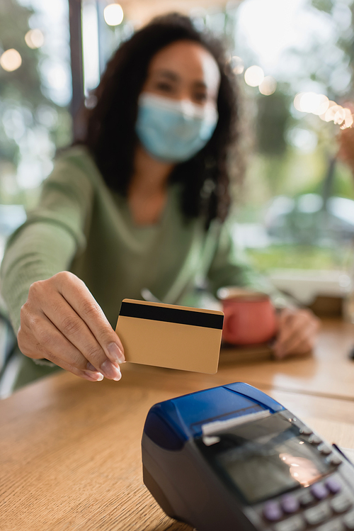 credit card near payment terminal in hand of african american woman in medical mask on blurred background