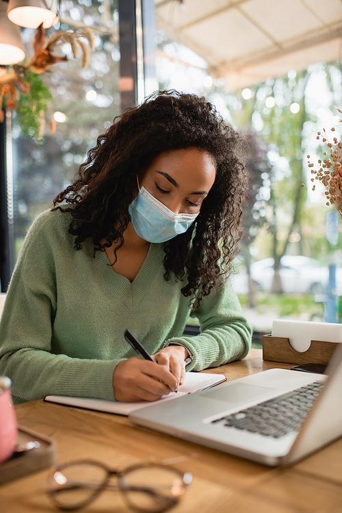 african american woman in medical mask writing in notebook near laptop in cafe